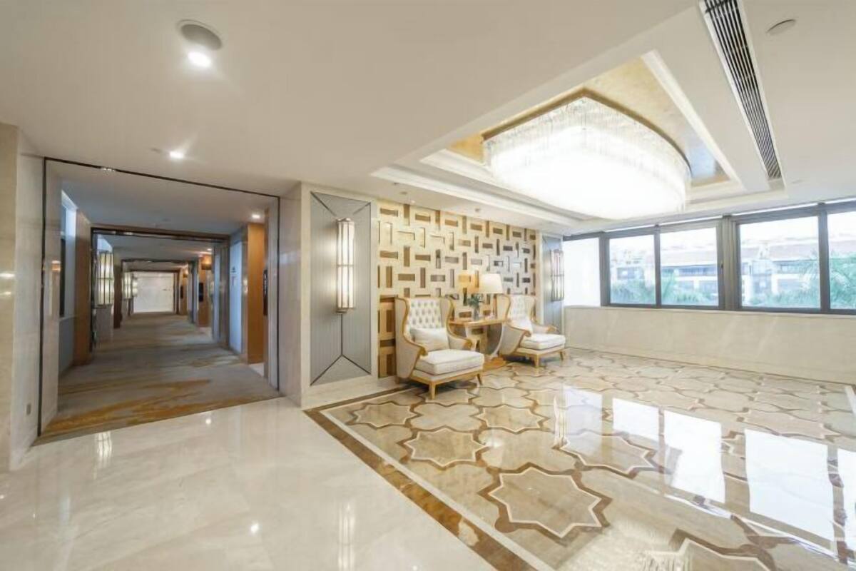 A spacious hotel lobby with a modern design, featuring a geometric patterned wall, a chandelier, and a large window offering a view.