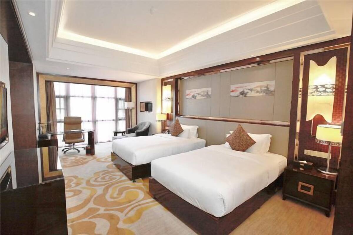 A hotel room with two beds, a desk, a chair, a large window, and a decorative mirror.