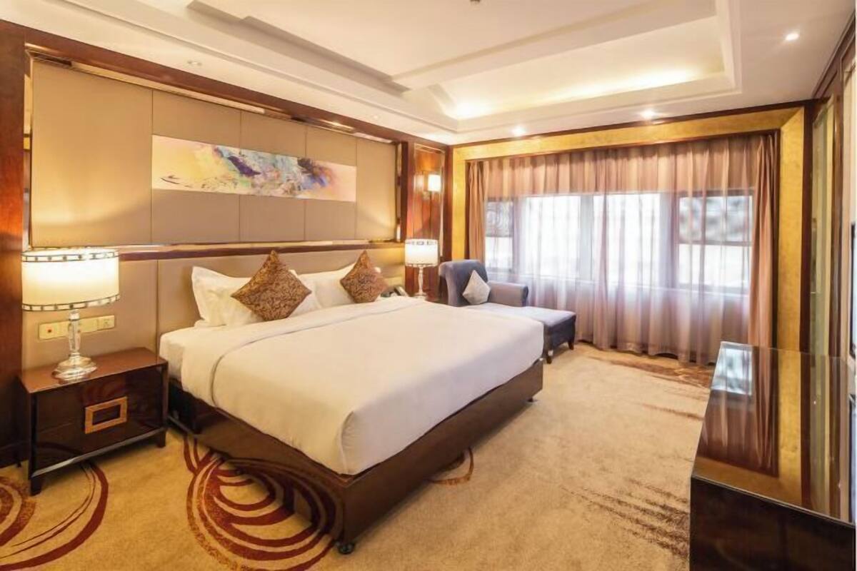 A spacious hotel room with a large bed, a bedside table with a lamp, a chair, and a large window with curtains.