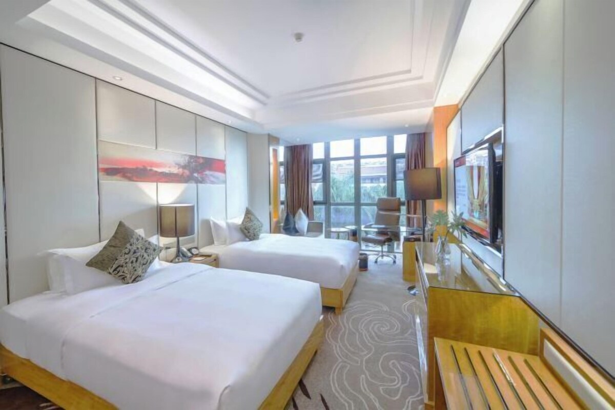 A hotel room with two beds, a desk, a chair, a TV, and a large window with curtains.