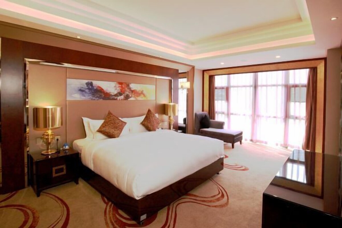 A spacious hotel room with a large bed, a nightstand, a desk, and a chair. There is a large window providing natural light.