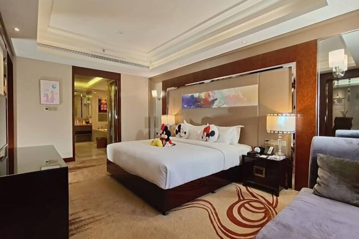 A hotel room with a large bed, a bedside table with a lamp, a TV, and a view of the hallway.