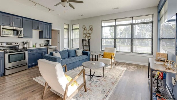 A modern kitchen with stainless steel appliances, a blue sofa, a white armchair, and a dining table with a rug.