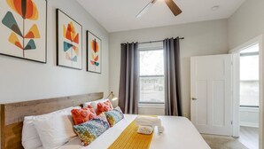 A hotel room with a bed, a ceiling fan, a window with curtains, and artwork on the wall.