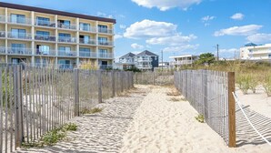 Ein sandiger Pfad führt zu einem mehrstöckigen Gebäude mit Balkonen, umgeben von einem Zaun und Strandpflanzen.