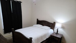 Un lit simple avec un cadre en bois et une literie blanche, une table de chevet avec une lampe, et un rideau noir à la fenêtre.