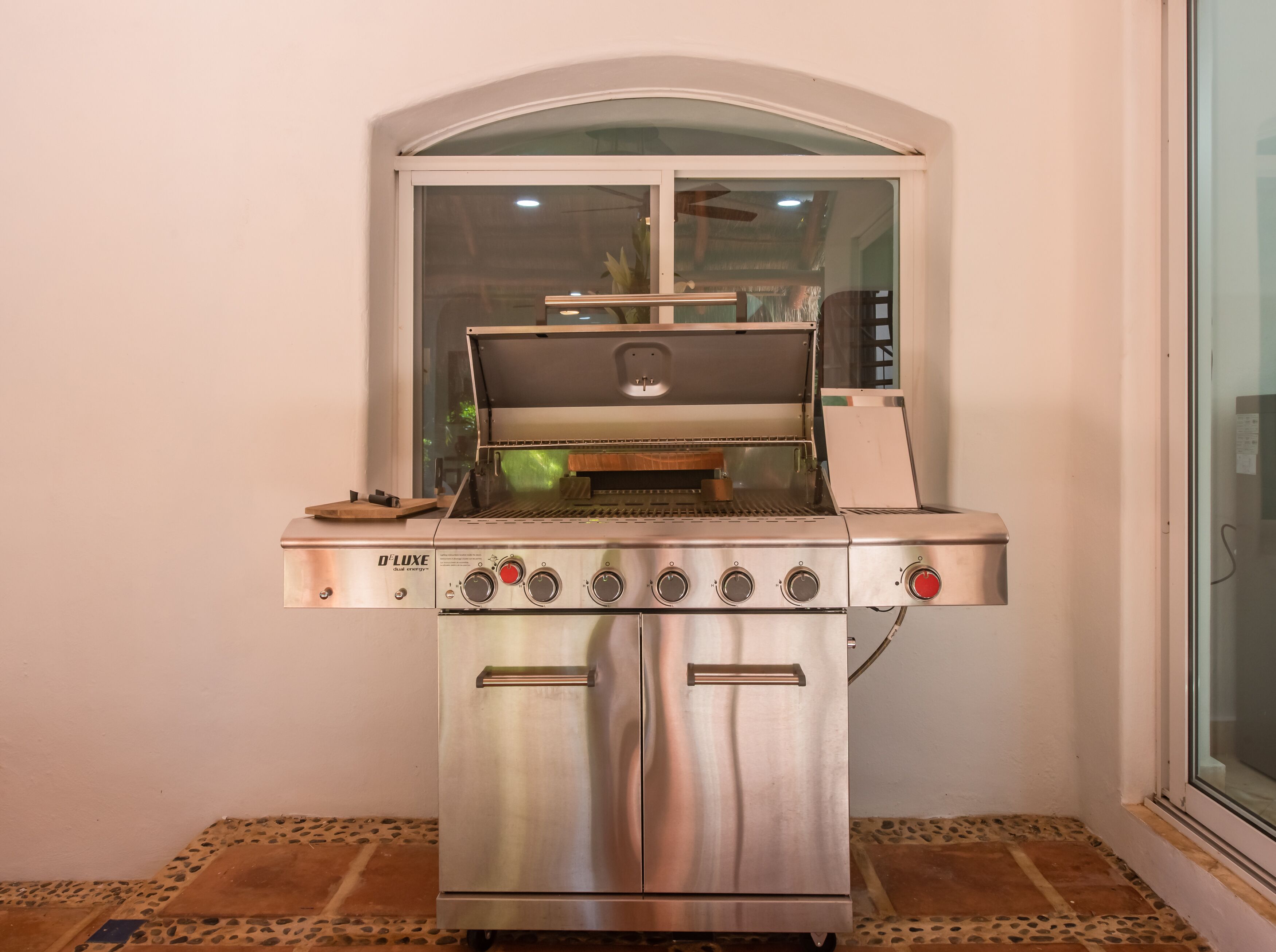 A stainless steel D'Lux dual energy grill with a window above it, positioned in a corner with tiled flooring.