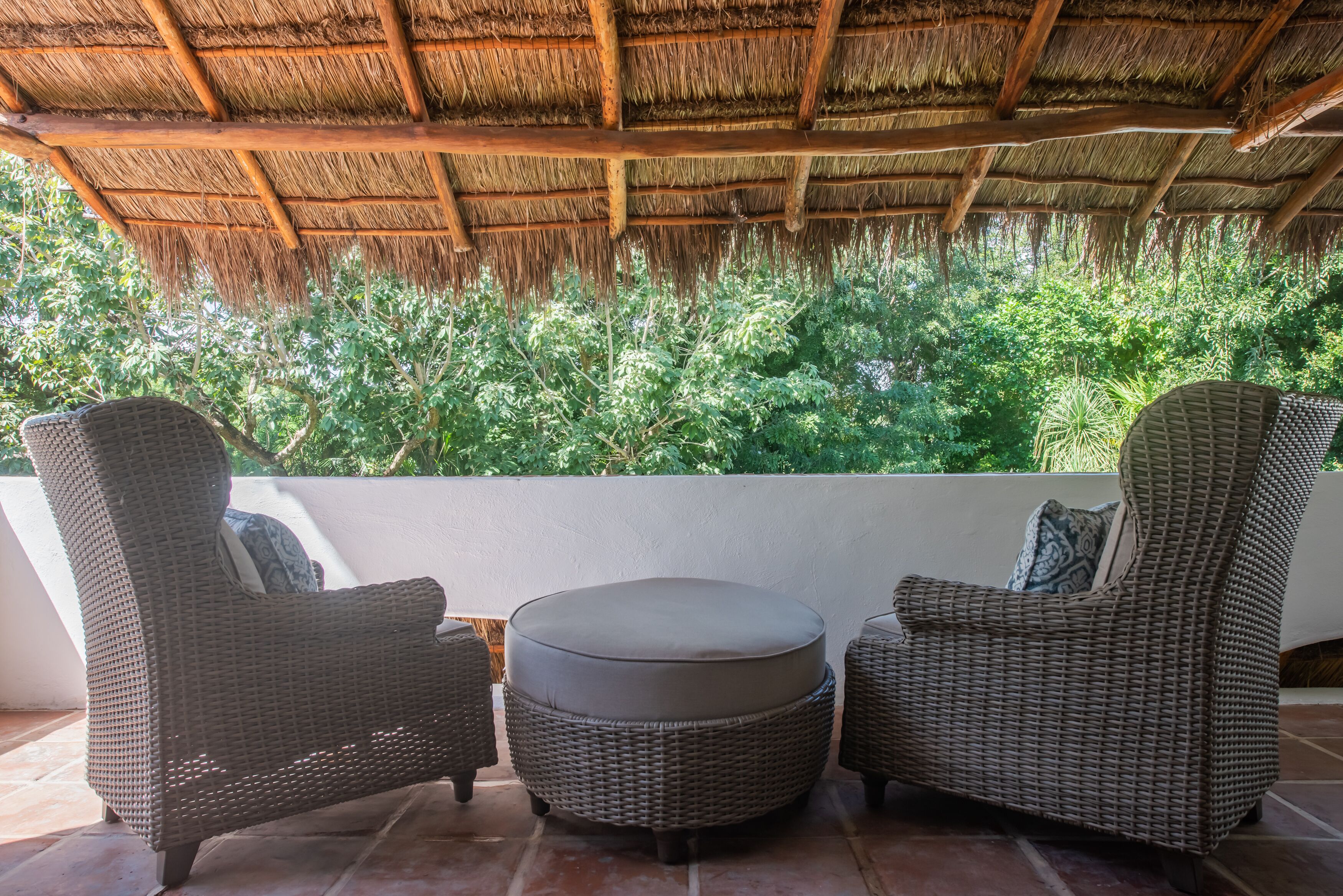 A patio with wicker furniture and a thatched roof, overlooking lush greenery.