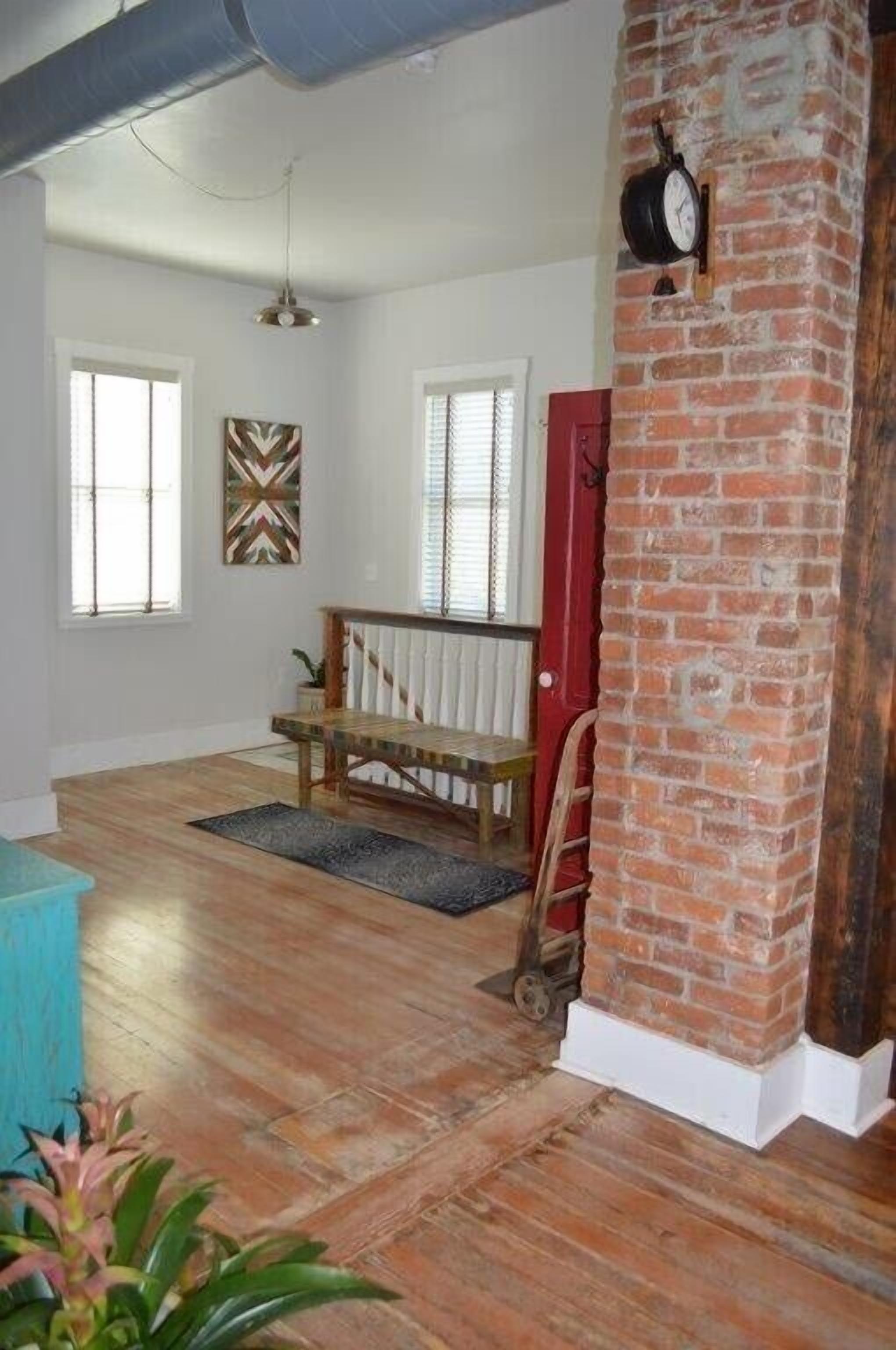 A spacious room with wooden flooring, a bench, a red door, and a brick column.
