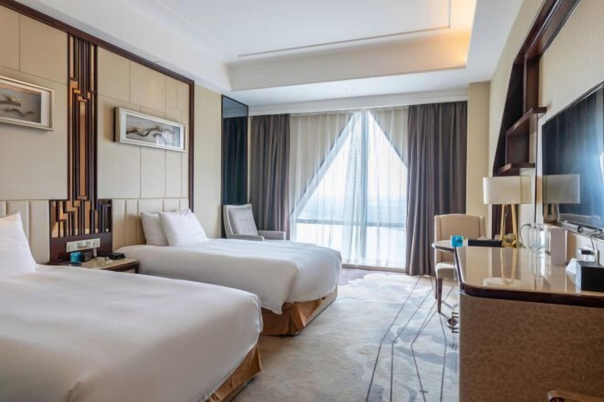 A hotel room with two beds, a desk, a chair, a TV, and a large window with curtains.