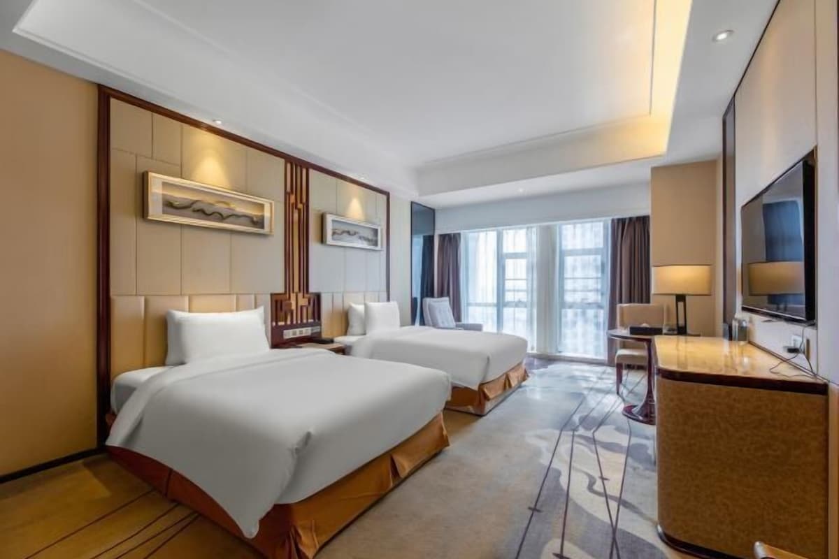 A hotel room with two beds, a desk, a TV, and a large window.