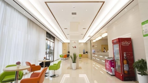 A modern hotel lobby with a reception desk, seating area with green and orange chairs, a vending machine, and a large potted plant.