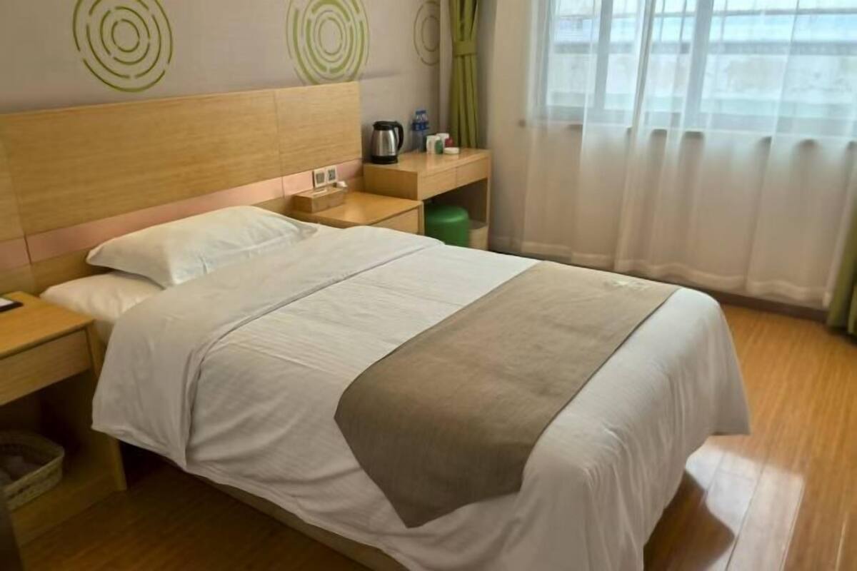 A hotel room with a wooden headboard, a bed with white bedding, a bedside table with a lamp, a green trash bin, and a window with sheer curtains.