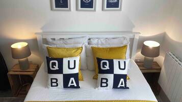 Una cama con cabecera blanca, dos lámparas de noche y almohadones decorativos que deletrean 'QUABA'.
