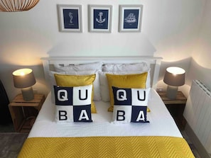 Una cama con cabecera blanca, dos lámparas de noche y almohadones decorativos que deletrean 'QUABA'.