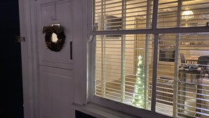 Ein Zimmer mit Fenster, ausgestattet mit horizontalen Jalousien, einer Tür mit einem Kranz und sichtbarem Innenraum, inklusive Essbereich mit Tisch und Stühlen.