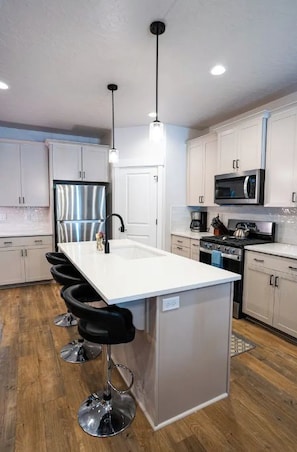 A modern kitchen with white cabinets, a stainless steel refrigerator, a microwave, and a bar area with stools. - Brand New 3 BR Vacation Home on a Golf Course (Victor)