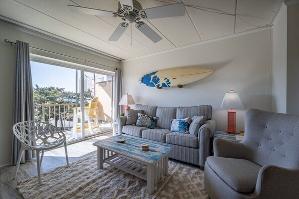 A living room with a grey sofa, a white chair, a coffee table, and a surfboard on the wall. - 1 Bedroom, Sleeps 4, Balcony w/ Ocean View (Ocean City)