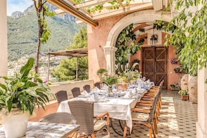 An outdoor dining area with a long table set for a meal, surrounded by wicker chairs, potted plants, and a view of a mountainous landscape. - Villa San Giacomo - Unique Villa in Positano (Positano)