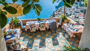 Une terrasse dotée d’un sol carrelé, de tables dressées pour les repas, et offrant une vue sur la mer et une ville côtière.
