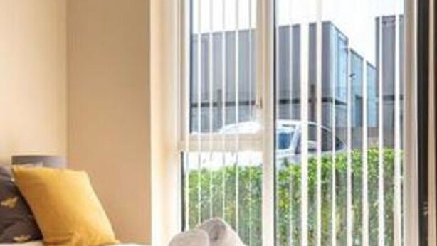 A room with a bed, a window with metal bars, and a view of a building and greenery outside.
