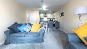 A modern living room with a grey sofa set, a dining table, and a kitchen in the background.