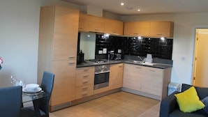 A compact kitchen with modern appliances, a dining area with a glass table, and a sofa with a yellow pillow. - Remarkable 2-bed Apartment in Reading (Reading)