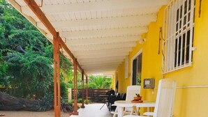 Un espacio al aire libre techado con sillas blancas y una mesa, paredes amarillas y piso de baldosas.