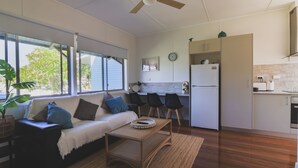 A modern living room with a sofa, coffee table, and chairs, a kitchen with a refrigerator, and large windows with a view of greenery. - RYDER FLATS UNIT 2 - Close to Beaches (South West Rocks)