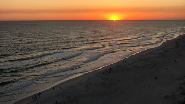 A beach at sunset with the sun near the horizon, calm sea, and people walking along the shore. - Beautiful beach condo for rent (Panama City beach)