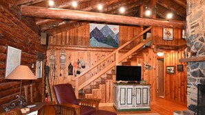 L’intérieur d’une cabane en rondins, avec un escalier en bois, un téléviseur, une cheminée et un coin salon confortable.