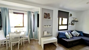 Un salón con piso de madera, muebles blancos, un sofá oscuro con cojines estampados y una ventana con cortinas.