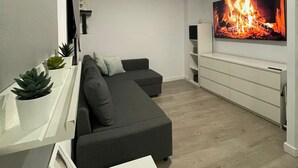 Una sala moderna con un sofá seccional gris, una mesa de centro blanca y un televisor de pantalla plana montado en la pared que muestra una imagen de una chimenea.