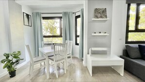 Una sala de estar moderna con una mesa de comedor blanca, dos sillas blancas, un sofá gris, una mesa de centro blanca y un sofá gris. Hay dos ventanas con cortinas, una planta en maceta y una estantería con objetos decorativos.