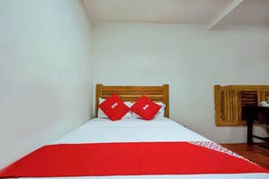 Una cama con cabecera de madera, dos almohadas rojas, una colcha blanca y una manta a rayas rojas y blancas.