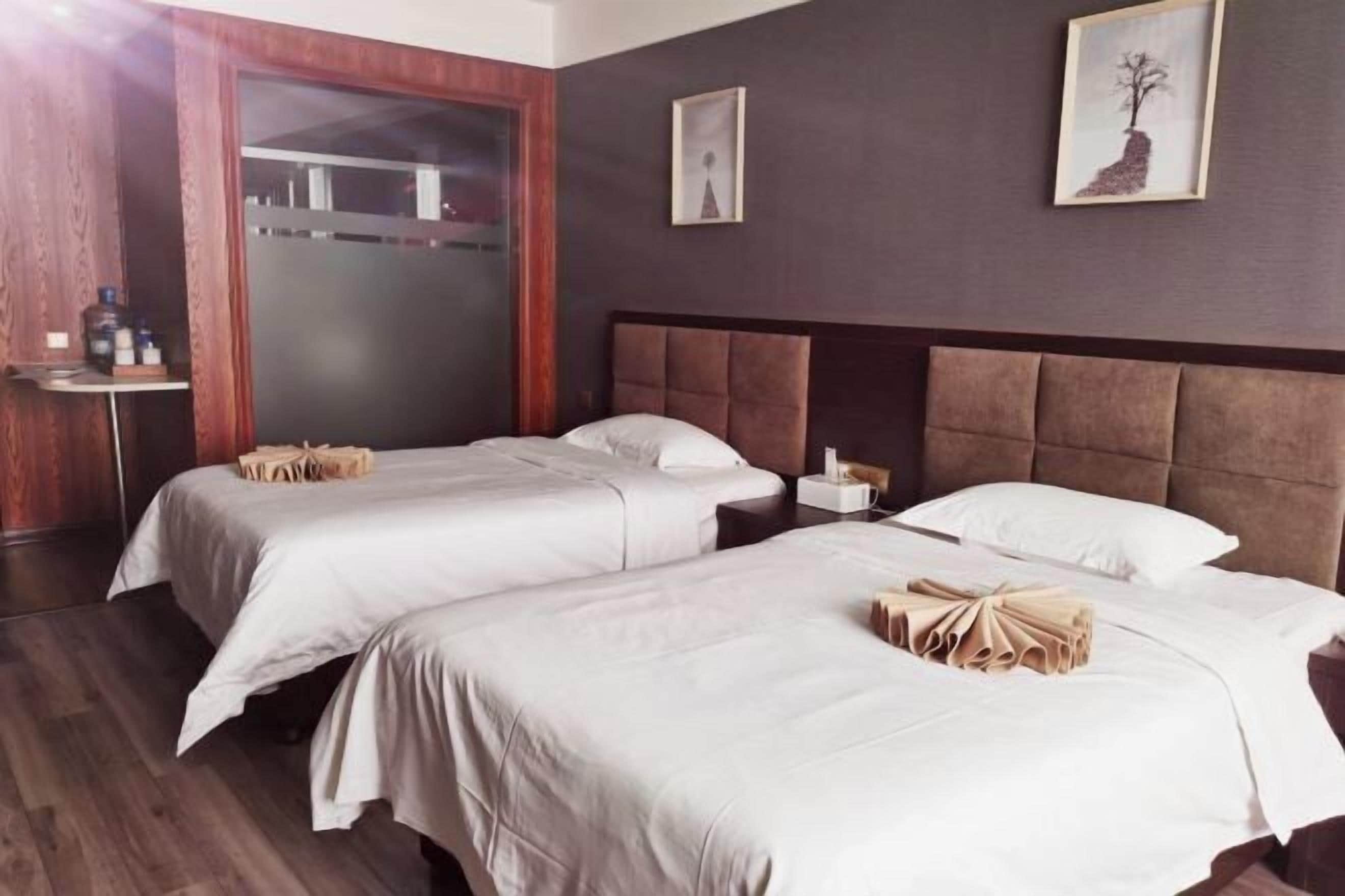 A hotel room with two beds, a wooden wardrobe, a bedside table, and a wall-mounted artwork.