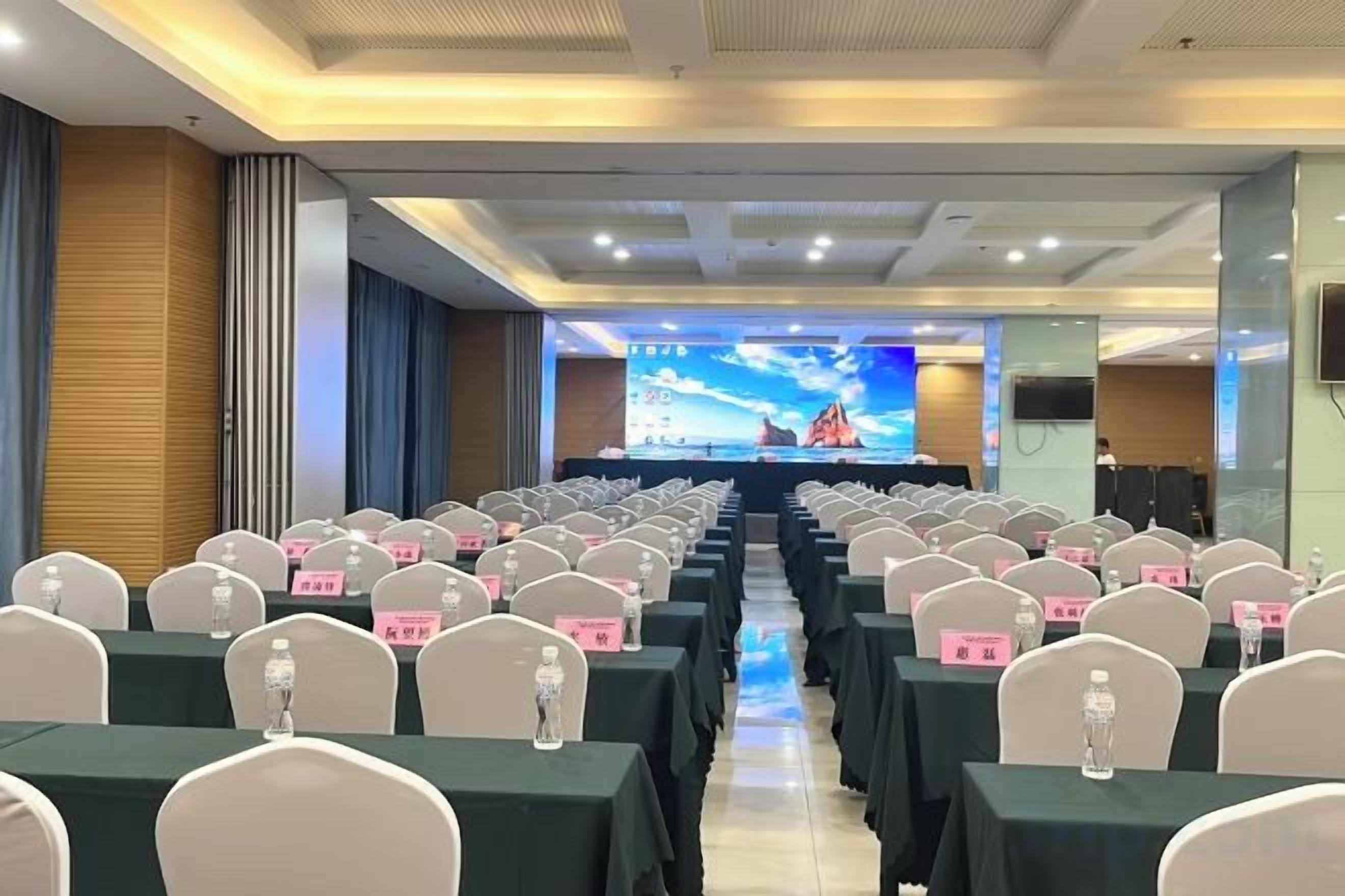 A conference room with rows of tables and chairs, a stage with a presentation screen, and a large window.