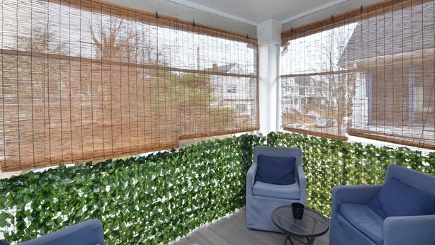 A covered patio area with two blue chairs and a small table, surrounded by green foliage and bamboo blinds.
