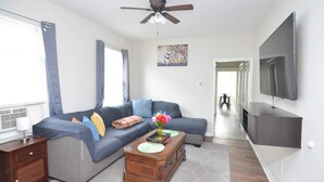 A modern living room with a sectional sofa, a wooden coffee table, a flat-screen TV mounted on the wall, and a ceiling fan.