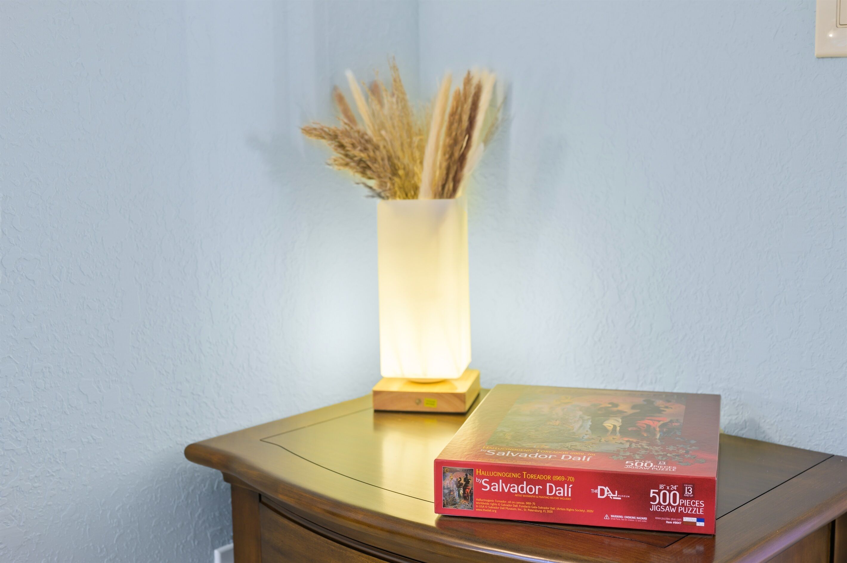Une boîte de puzzle sur une table en bois, avec un vase contenant des plantes séchées et une lampe allumée.