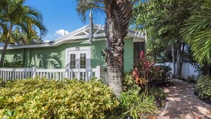 A green building with a white fence, a palm tree, and various plants.