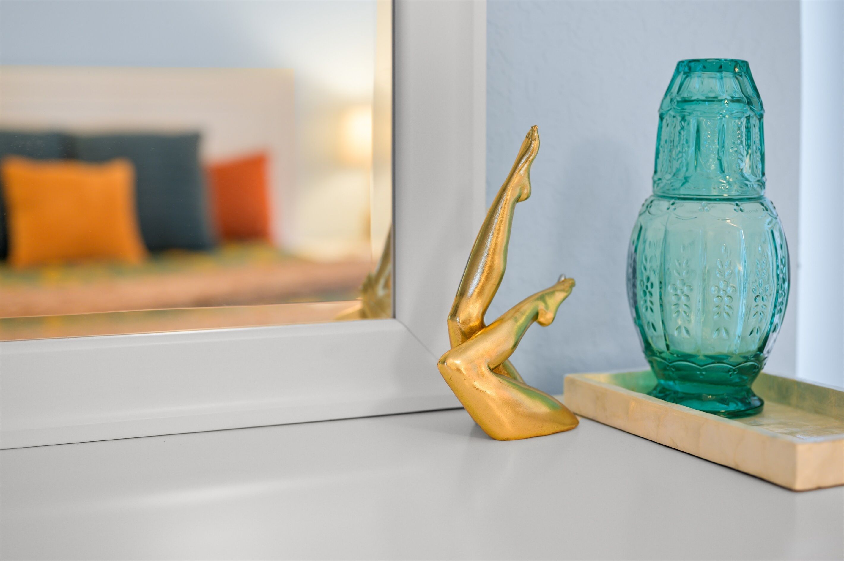 A decorative glass vase on a wooden tray, a golden sculpture, and a white-framed mirror reflecting a bed with pillows.