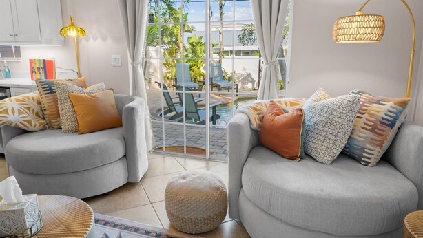 A modern living room with two grey sofas, patterned cushions, a small round ottoman, and a dining area with a table and chairs.