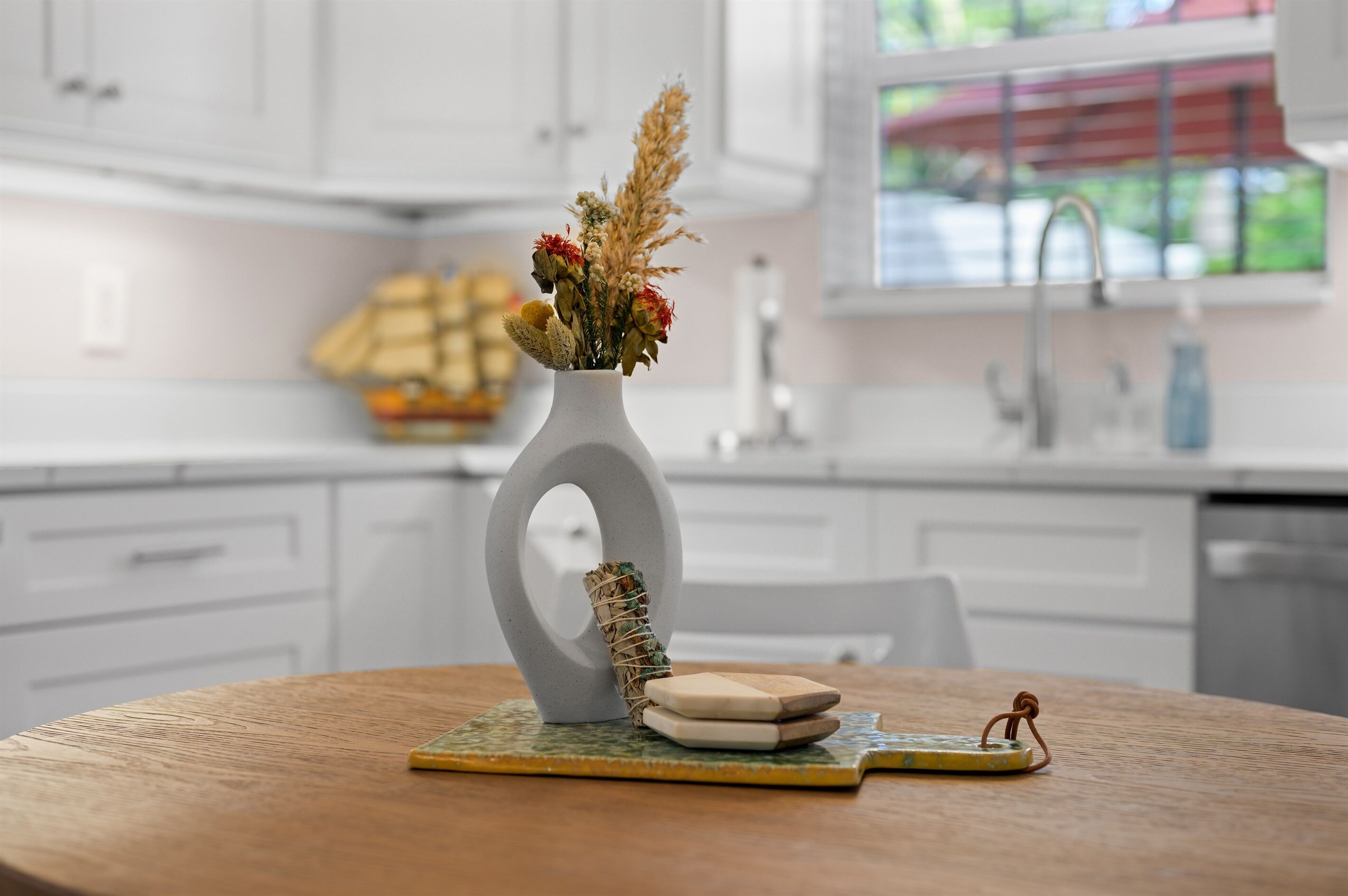 Une cuisine moderne avec des meubles blancs, un vase contenant des fleurs séchées et une table en bois sur laquelle se trouvent un dessous de verre et une petite figurine.