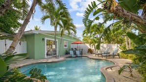 A swimming pool with lounge chairs and umbrellas, surrounded by tropical plants and a green house.