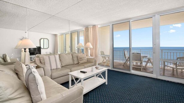 A spacious living room with a large sofa, a coffee table, and a TV. There are two lamps and a window with a view of the ocean. - Oceanfront 2 Bed/2 Bath, Sleeps 6, Pools, Gym, Etc (Ocean City)