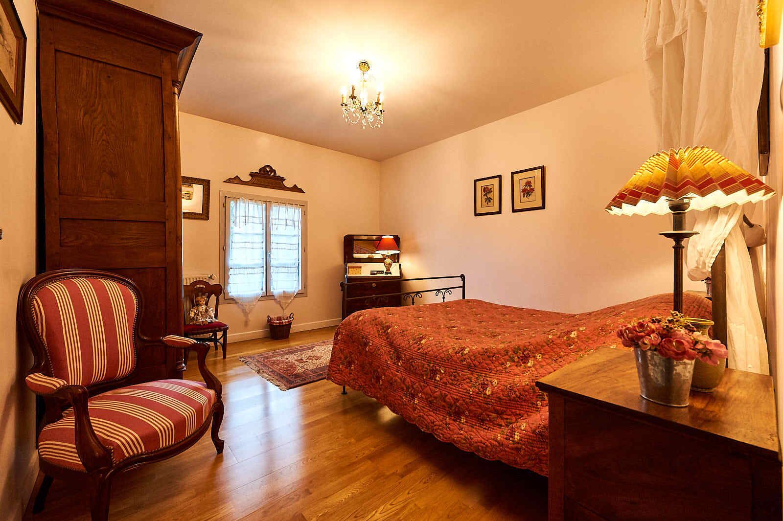 A bedroom with a wooden bed, a chair, a dresser, a nightstand, a lamp, and a window with curtains.