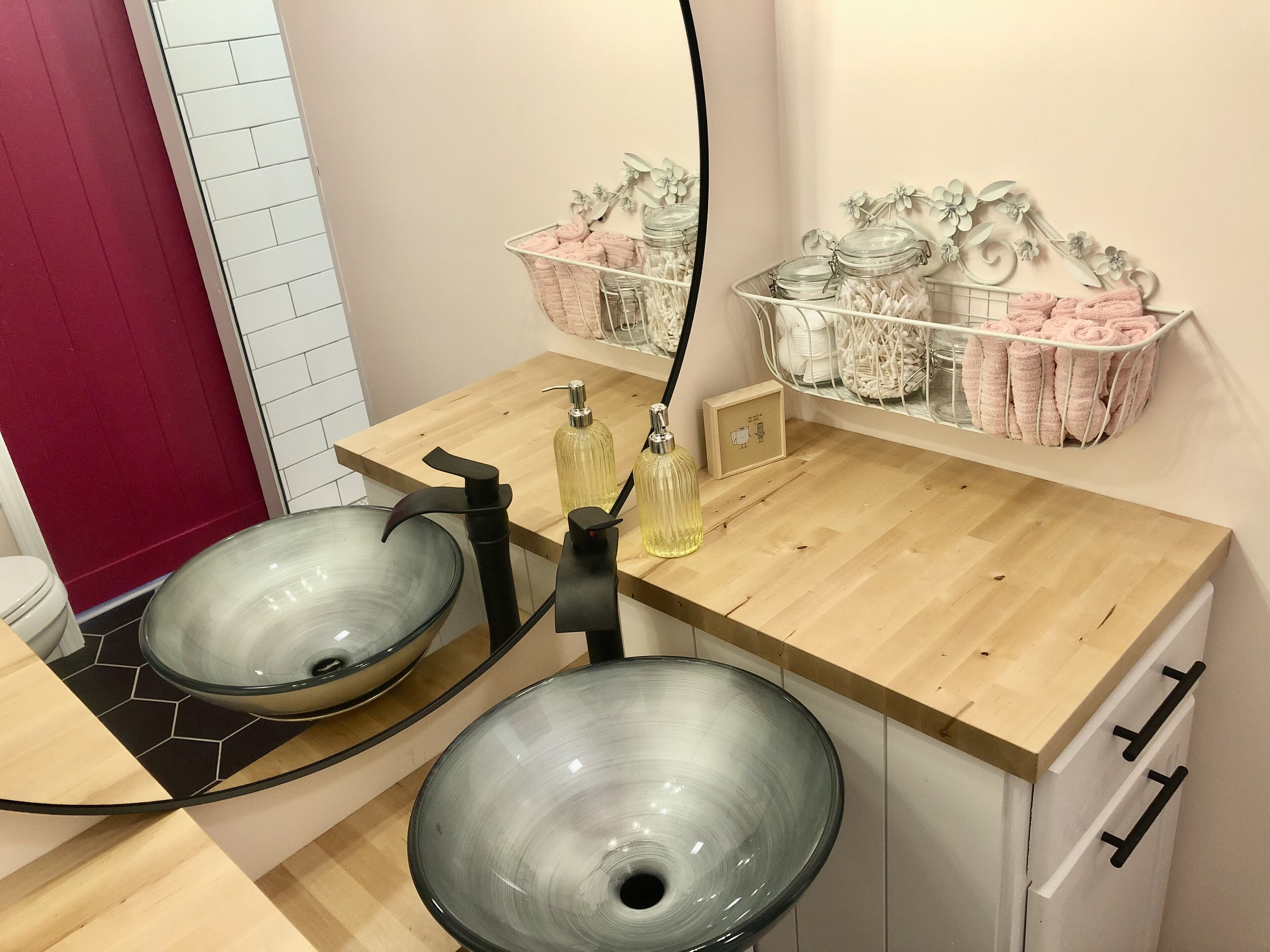 A bathroom with two glass sinks, a wooden countertop, and a mirror. There are two metal baskets on the wall, one with pink towels and the other with jars.