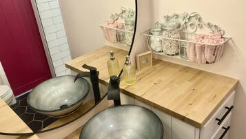 A bathroom with two glass sinks, a wooden countertop, and a mirror. There are two metal baskets on the wall, one with pink towels and the other with jars.