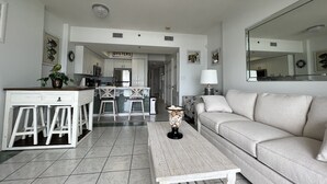 A modern living room with a sofa, coffee table, and kitchen in the background. - Newly Remodeled, Beachfront Condo, Sleeps Six, Resort Amenities! (Orange Beach)